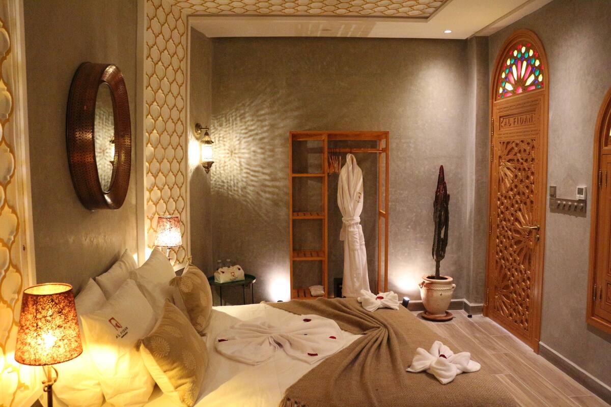 A hotel room with a bed, a mirror, a wall-mounted light, a wooden door, and a potted plant.
