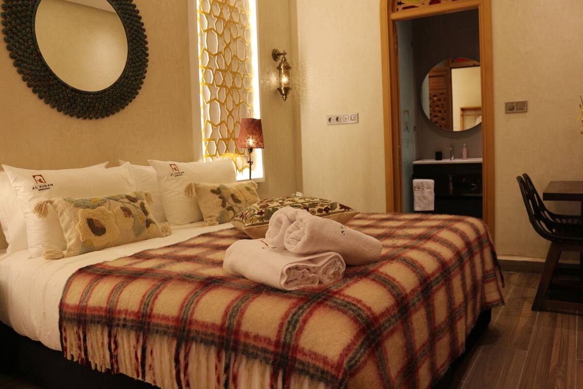 A neatly made bed with a plaid blanket, a bedside lamp, and a wall-mounted mirror.