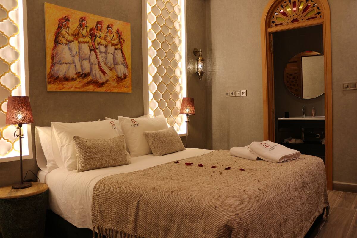A neatly made bed with a beige blanket and pillows, a painting on the wall, and a lamp on the side table.
