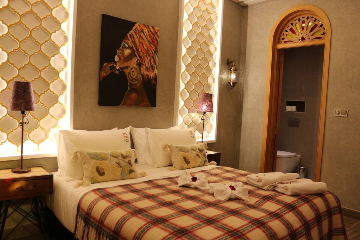 A hotel room with a bed, a painting, a lamp, and a bathroom door.