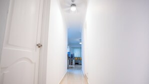 A narrow hallway with a door on the left, a ceiling light, and a glimpse of a living area with a sofa and a table in the distance.