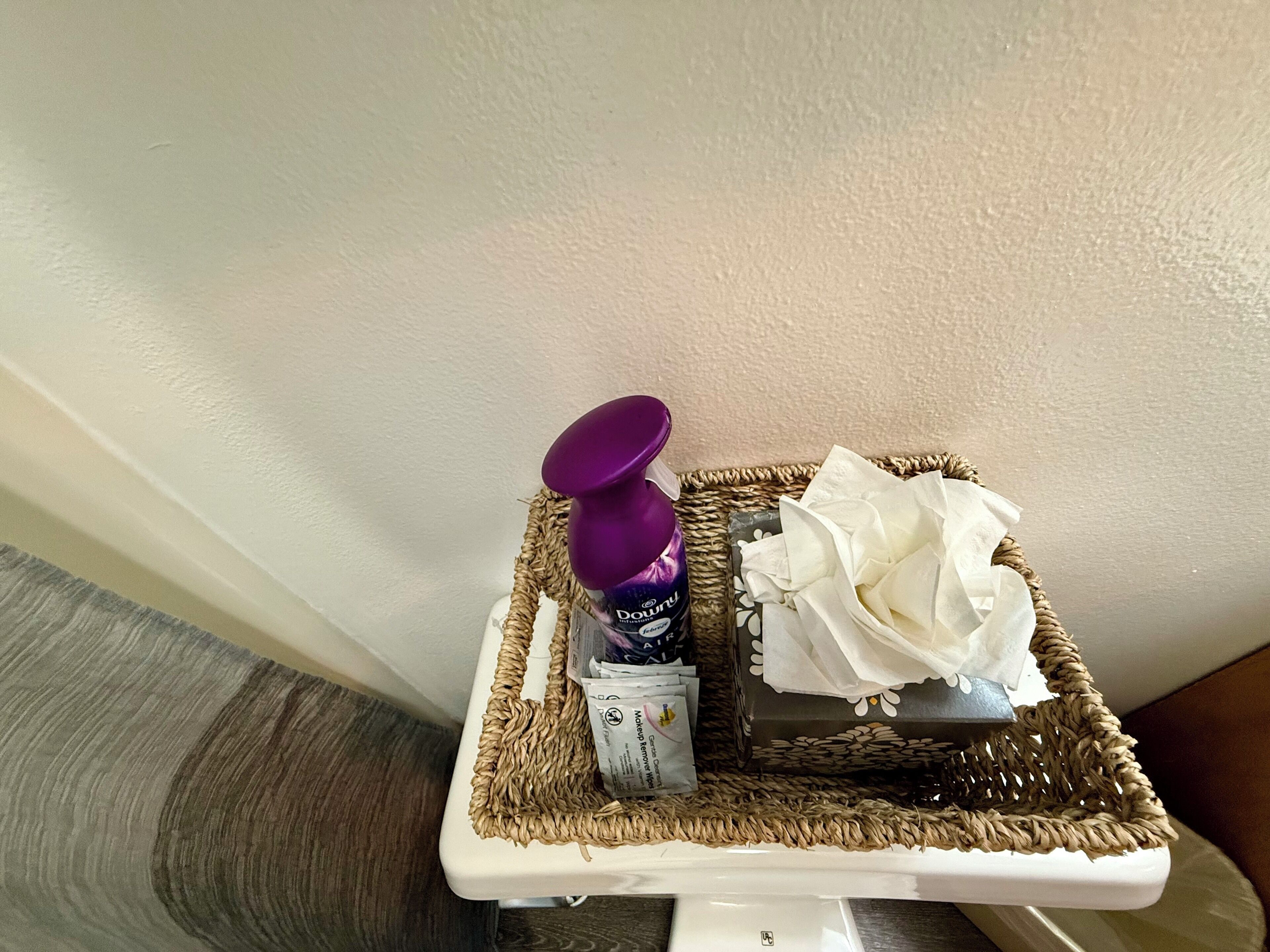 A bathroom corner with a white toilet, a wicker basket containing a purple spray bottle, a tissue box, and a tissue.