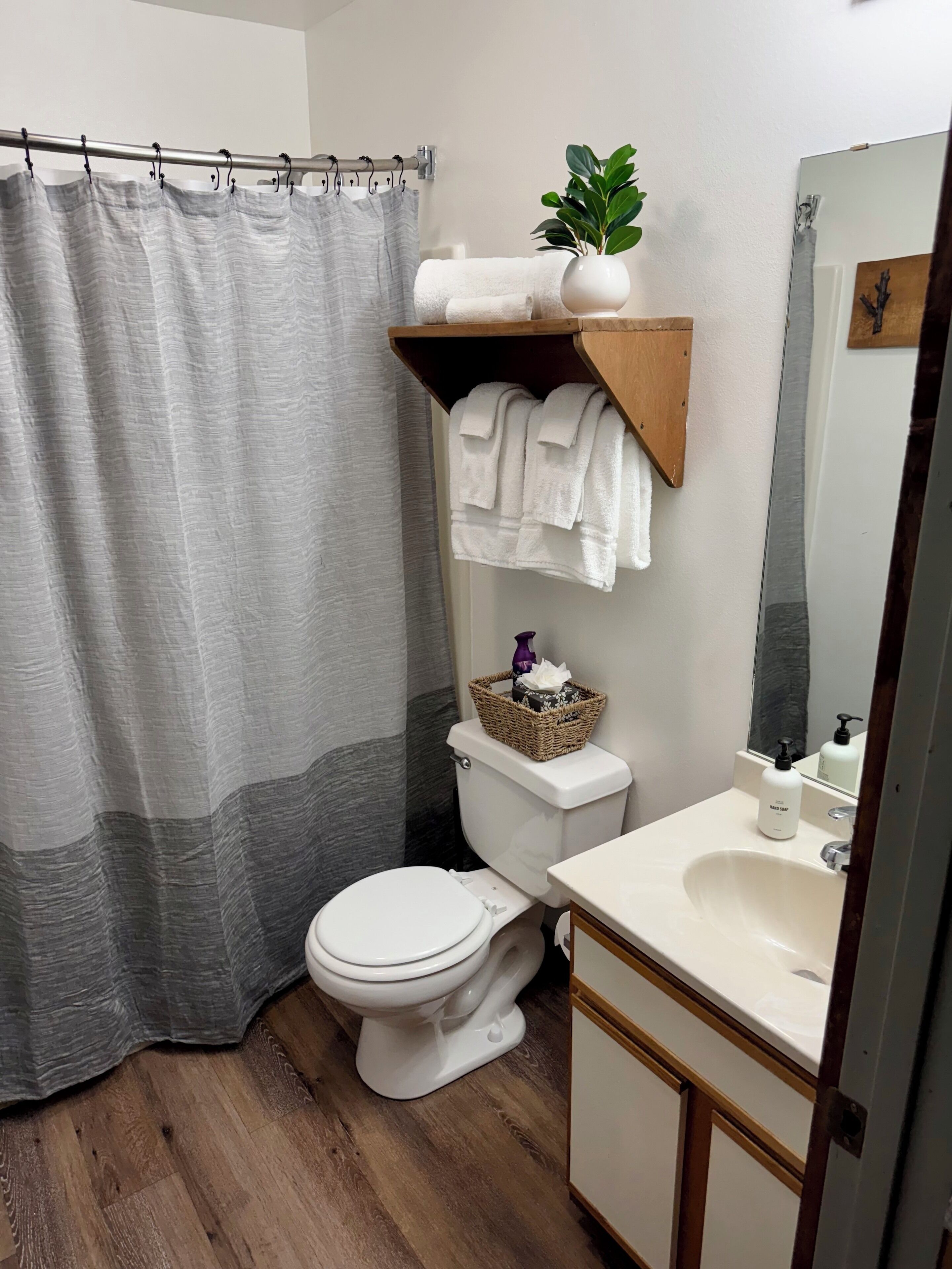 A bathroom with a shower curtain, a toilet, a sink, a mirror, and a shelf with towels and a plant.