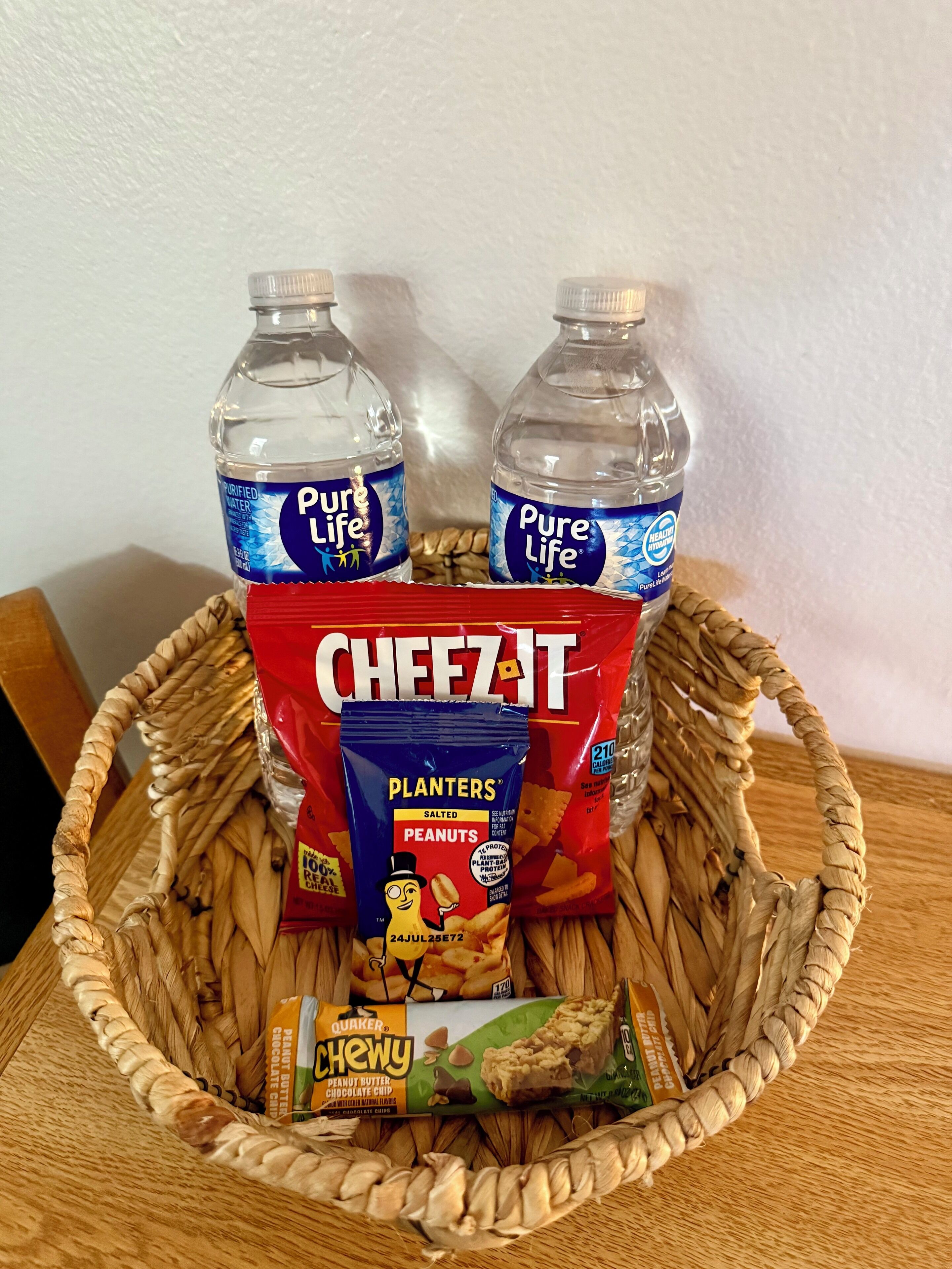 A basket containing two bottles of Pure Life water, a bag of Cheez-It, a bag of Planters salted peanuts, and a Quaker Chewy peanut butter chocolate chip bar.