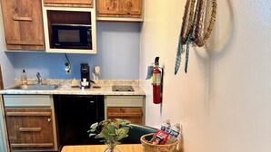 A compact kitchen with wooden cabinets, a microwave, a sink, and a fire extinguisher. There's a table with a vase and a basket containing bottled water and a snack.