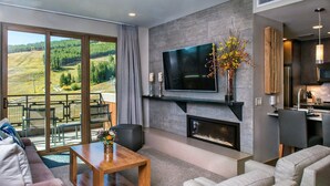 Una sala de estar moderna con chimenea, un televisor de pantalla plana grande y vistas a un paisaje boscoso a través de amplios ventanales corredizos.
