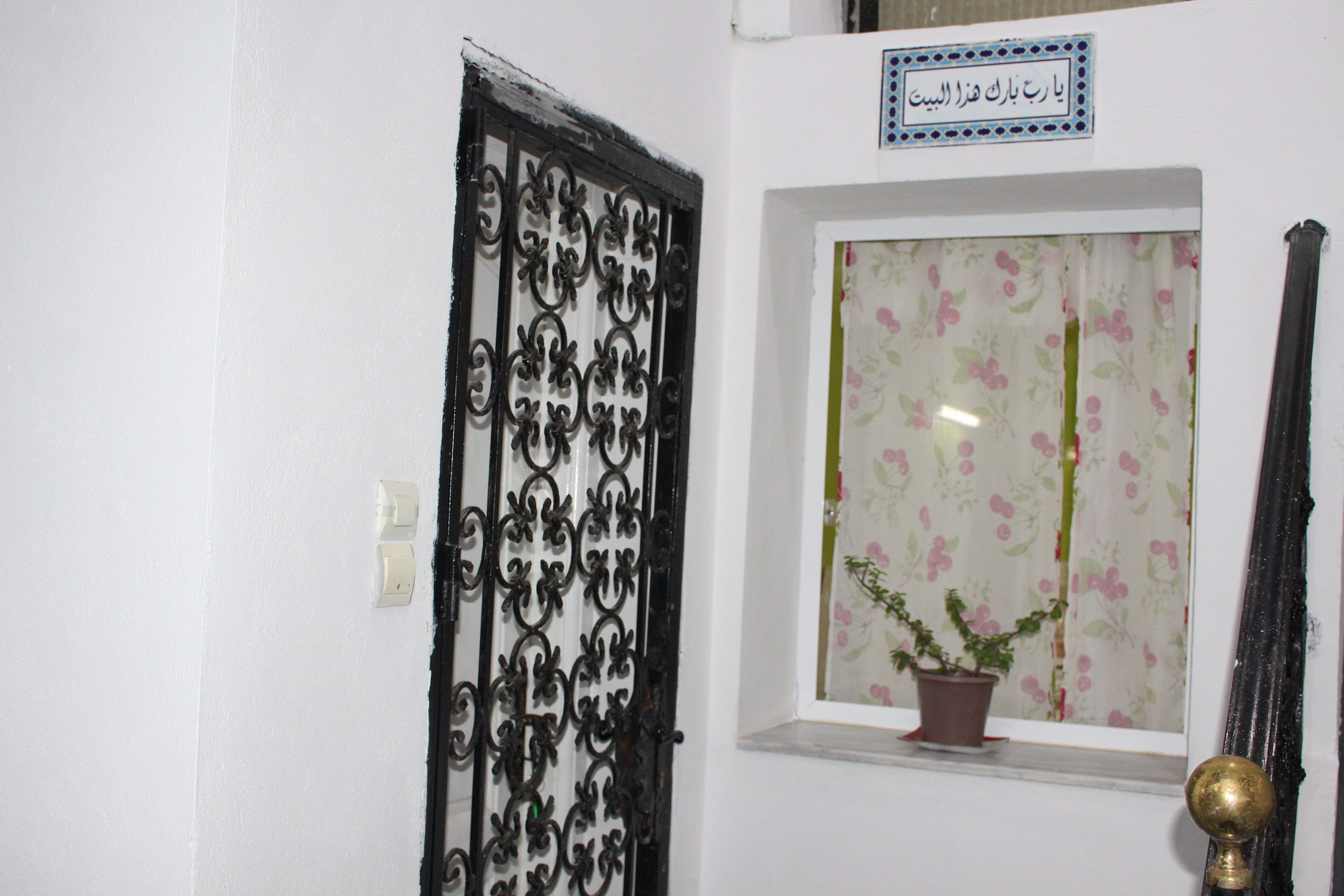 Une porte ornée de motifs complexes en métal noir, un pot de fleurs posé sur un rebord de fenêtre et une enseigne avec du texte en arabe.