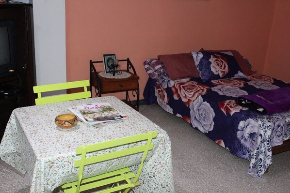 A room with a bed covered in a floral duvet, a table with a magazine, and a chair.