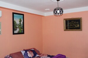 A bedroom with a bed, a framed picture of a landscape, and a framed Arabic calligraphy piece on the wall.