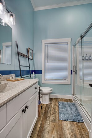 Un baño con piso de madera, cabina de ducha de vidrio, inodoro e lavamanos con espejo arriba.