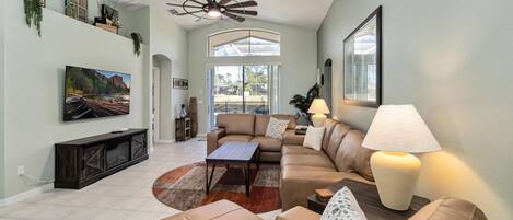 A spacious living room with a large sectional sofa, a coffee table, and a TV mounted on the wall. There is a ceiling fan, a fireplace, and a large window providing natural light.