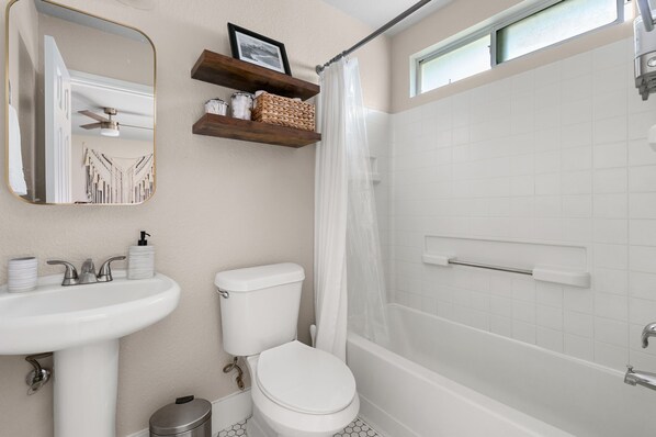 A bathroom with a white sink, toilet, and bathtub. There is a mirror above the sink, a shower curtain, and a window above the bathtub.