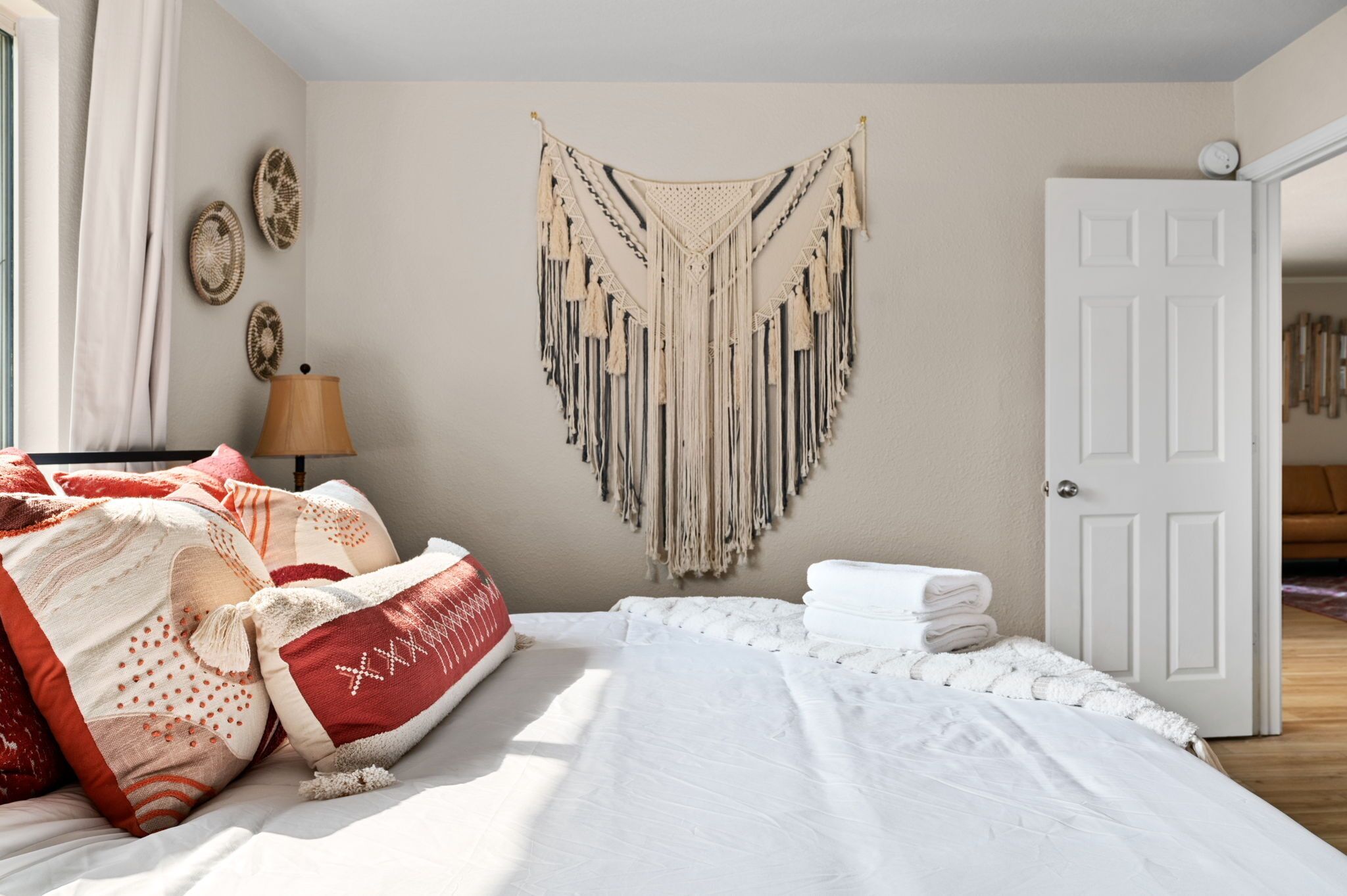 A neatly made bed with white and red bedding, a macrame wall hanging, and a white door in a room with beige walls.