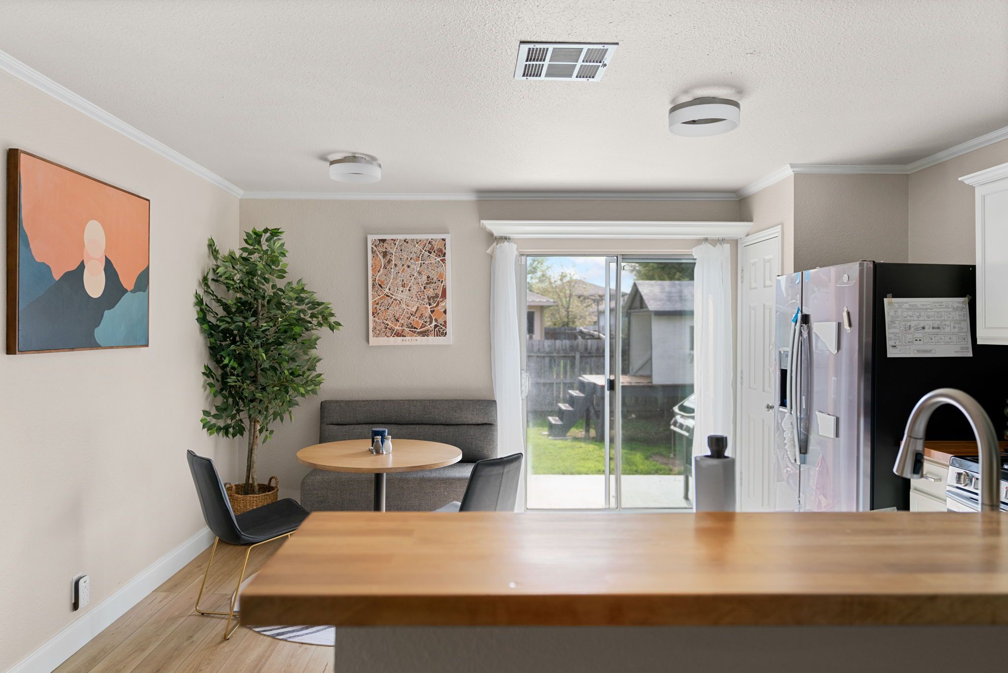A modern kitchen with a wooden countertop, a dining area with a round table and chairs, a potted plant, and a sliding glass door leading to a backyard.