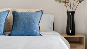 A neatly made bed with blue and beige pillows, a white comforter, and a black vase with a plant on a wooden nightstand. - Solace in South Austin - Game Room (Austin)