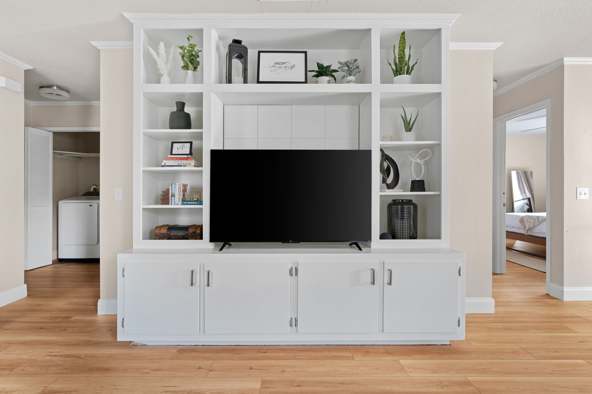 A modern living room with a large flat-screen TV on a white entertainment unit, surrounded by shelves with decorative items and plants.