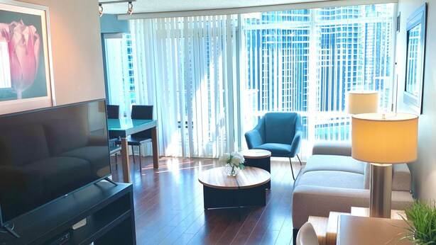 Una sala de estar moderna con un televisor de pantalla plana, un sofá, una mesa de centro y una silla. Hay un ventanal con persianas y vistas a edificios altos.