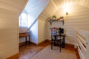 A room with wooden walls, a sloped ceiling, a window, a table, a chair, a rug, and a shelf with books and a quilt.