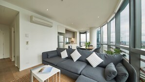 Een moderne woonkamer met een grijze bank, een klein wit bijzettafeltje en een uitzicht op de stad door grote ramen.