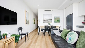 Una sala de estar moderna con un sofá de cuero negro, un comedor con mesa y sillas de madera, un televisor de pantalla plana montado en la pared, y una cocina con refrigerador y lavavajillas.