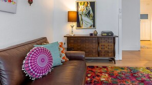 Un salón con un sofá de cuero marrón, cojines de colores, un aparador de madera, una lámpara y un cuadro en la pared.