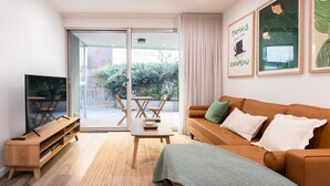 Un salon moderne avec un canapé, une table basse et un téléviseur posé sur un meuble en bois. Une porte-fenêtre coulissante donne accès à un balcon équipé d’une table et de chaises.