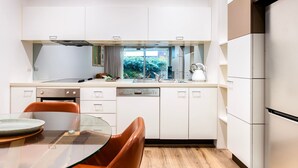 Une cuisine moderne avec des meubles blancs, une table à manger en verre et une vue sur la verdure à l’extérieur.