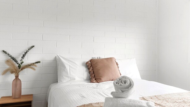 A neatly made bed with white bedding and a single brown pillow, a small wooden nightstand with a vase of dried plants, and a rolled white towel on the bed. - Rosebud Beachfront Haven  Coastal Bliss Four (Capel Sound)