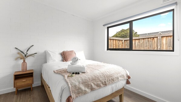 A bedroom with a white brick wall, a wooden bed frame, a white bedspread, a pink pillow, a white vase with dried plants, and a window with a view of a wooden fence.
