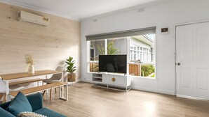 A modern living room with a wooden floor, a white door, a TV on a white stand, a dining table with chairs, and a sofa. - Rosebud Beachfront Haven  Coastal Bliss Four (Capel Sound)