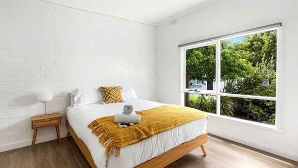 A bedroom with a large window, white brick walls, a wooden bed, and a bedside table with a lamp. - Rosebud Beachfront Haven  Coastal Bliss Five (Capel Sound)