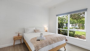 Quarto com uma janela grande, uma cama com manta estampada, mesa de cabeceira com abajur e vista para um jardim.