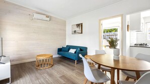 A modern living room with a blue sofa, a round wooden dining table, and a kitchen visible through an open door.