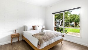 A bedroom with a large window, a wooden bed frame, white bedding, and a bedside table with a lamp.