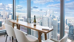A high-rise room with a city view, a wooden table, two chairs, a bottle of champagne, and a vase of dried flowers. - Sky High Serenity  Level 66, Docklands, Melbourne (Melbourne)