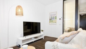Un soggiorno moderno con una TV a schermo piatto su un supporto bianco, una lampada a sospensione in vimini, un quadro di Van Gogh incorniciato e un divano bianco con una coperta decorata.