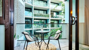 Un balcone moderno con ringhiera in vetro, due sedie e un tavolino, con vista su un edificio a più piani con balconi e vegetazione.