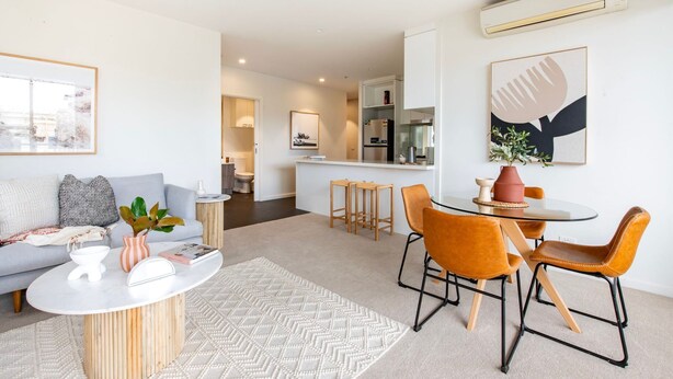 A modern living room with a grey sofa, a coffee table, and orange chairs. There is a kitchen area in the background with a fridge and a sink.