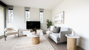 A modern living room with a large flat-screen TV, a grey sofa, a white armchair, and two round wooden side tables.