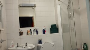 Ein Badezimmer mit weißem Waschbecken, WC und Dusche. Auf dem Waschbecken stehen Pflegeprodukte und ein Mülleimer steht daneben.