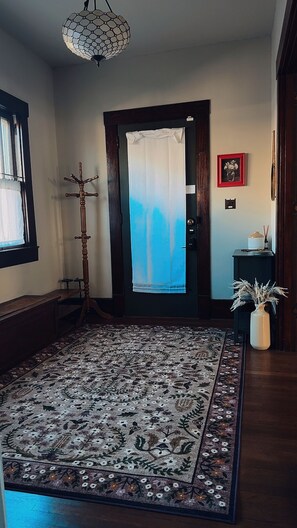 A hallway with a patterned rug, a coat rack, a door with a curtain, a framed picture, and a vase with dried plants.