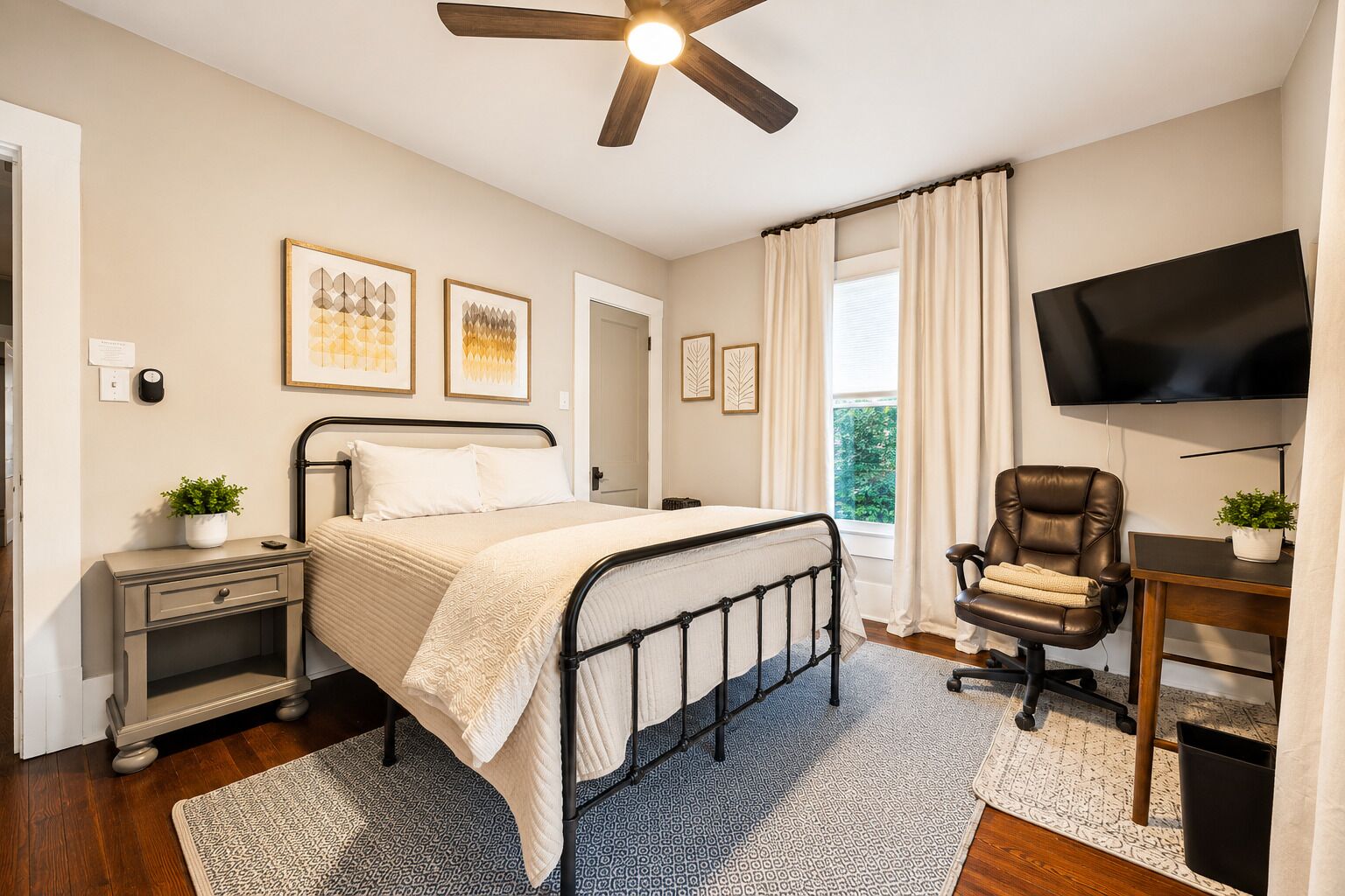 Une chambre à coucher comprenant un lit, une table de chevet, une chaise, un téléviseur, un ventilateur de plafond et des œuvres d’art accrochées au mur.