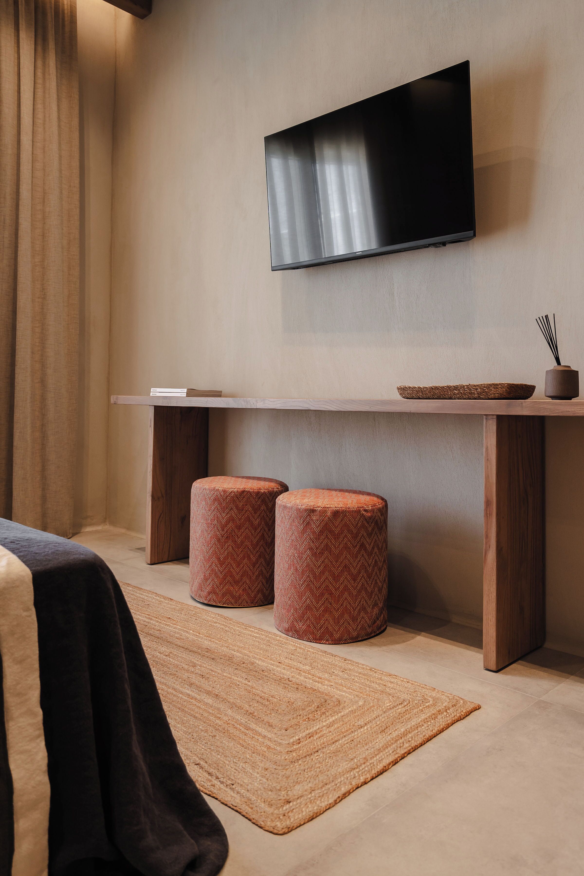 A modern hotel room with a flat-screen TV mounted on the wall, a wooden table with a woven tray, two ottomans, and a woven rug on the floor.