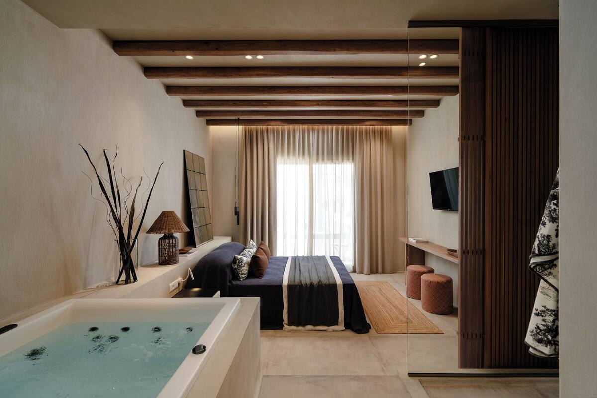 A modern bedroom with a large bathtub, a bed with a dark bedspread, a wooden ceiling, and a large window with curtains.