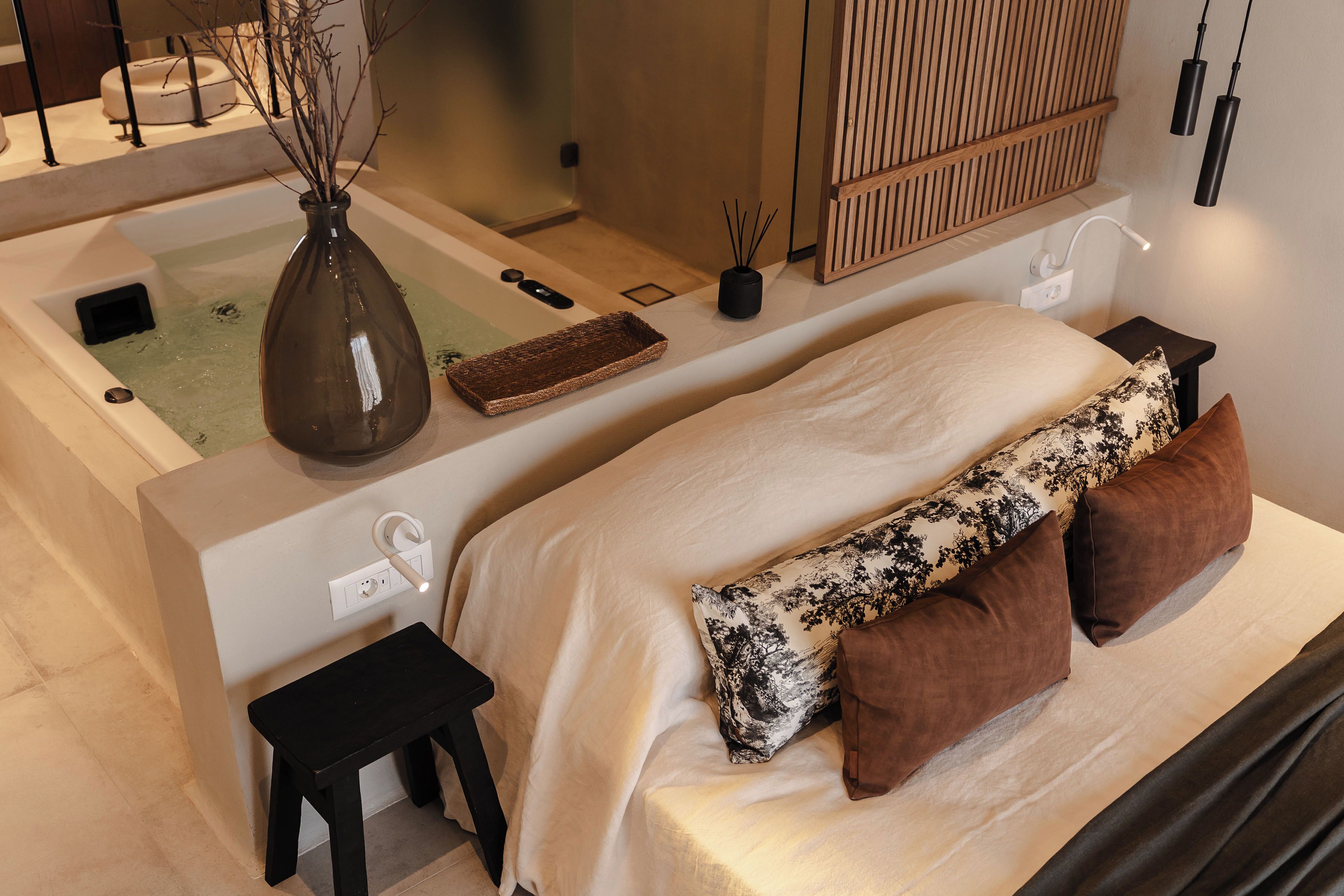 A modern spa room with a large bathtub, a small wooden stool, a black vase with branches, and a black incense burner.