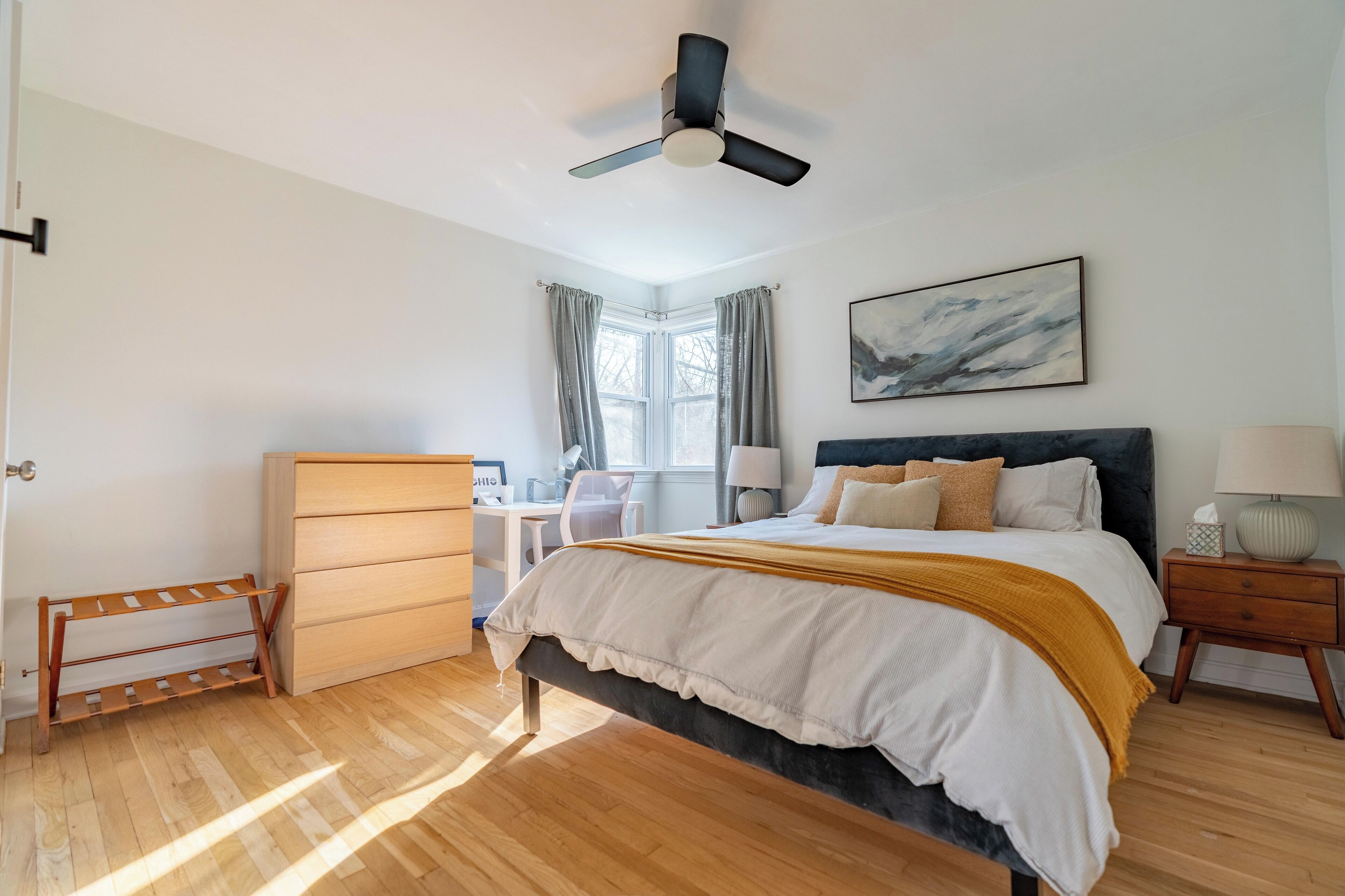 A bedroom with a bed, a desk, a chair, a wardrobe, a ceiling fan, and a window with curtains.