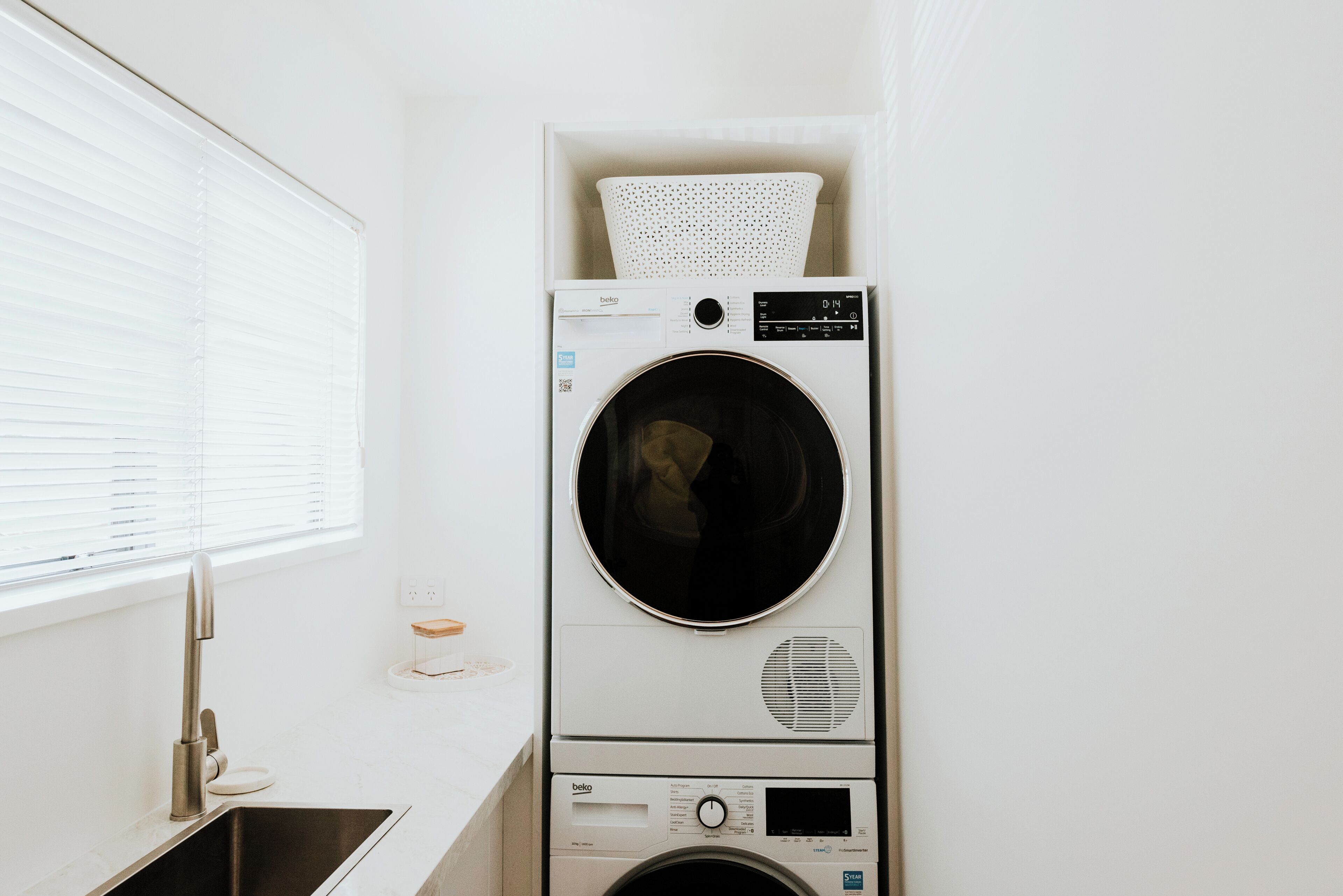 A compact laundry area with a stacked washer and dryer, a window with blinds, and a stainless steel sink.
