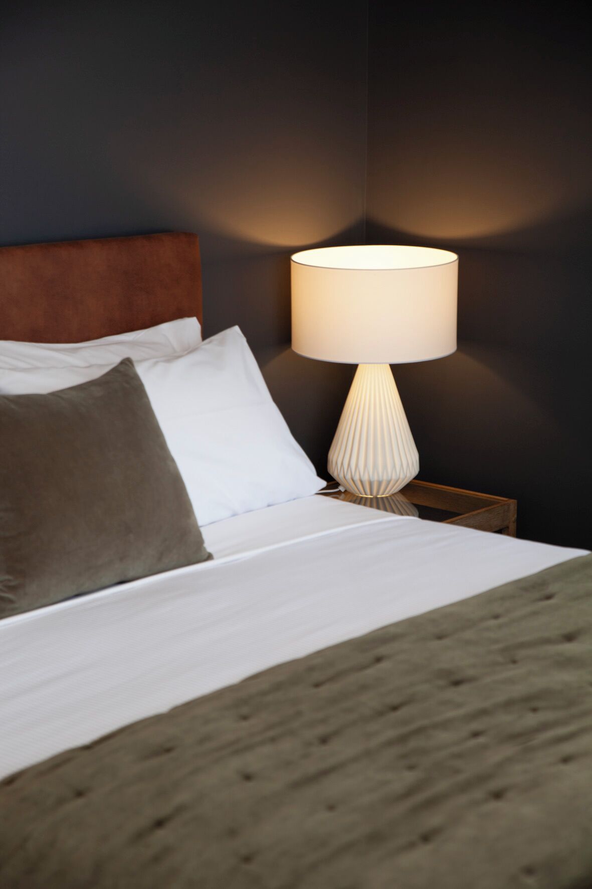 Una cama bien hecha con ropa de cama blanca y gris, un cabecero de madera y una lámpara blanca con base texturizada sobre una mesita de noche.