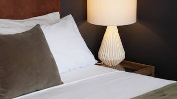 Una cama bien hecha con ropa de cama blanca y gris, un cabecero de madera y una lámpara blanca con base texturizada sobre una mesita de noche.