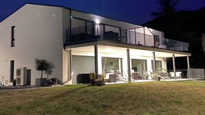 Une maison moderne à deux étages, dotée d’un balcon, d’une terrasse couverte et d’une pelouse.